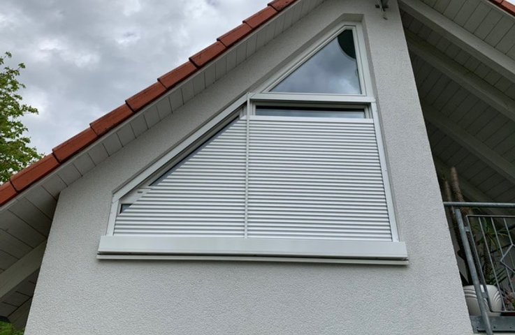 „Šikmé venkovní žaluzie na moderním rodinném domě – lomená fasáda se štítovým prosklením a zimní zahrada jsou zastíněné antracitovými žaluziemi, které kopírují sklon střechy