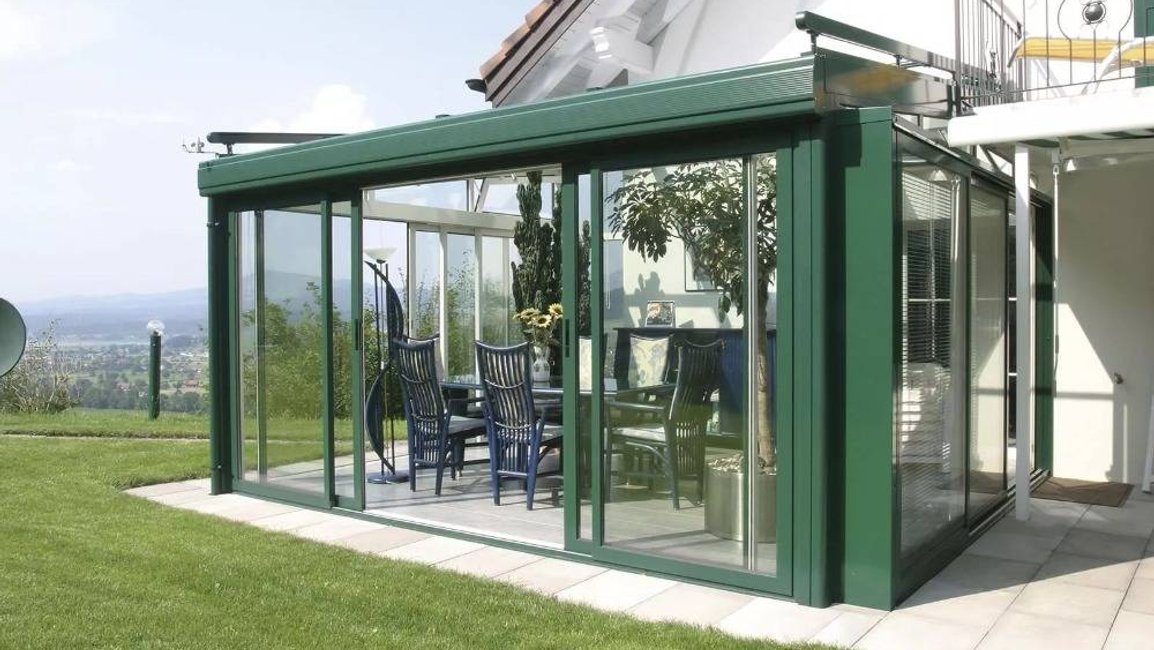 Moderní celoroční zimní zahrada v zelené hliníkové konstrukci s velkými posuvnými skleněnými stěnami vytváří u rodinného domu pohodlné místo pro posezení s výhledem do zahrady a okolní krajiny.