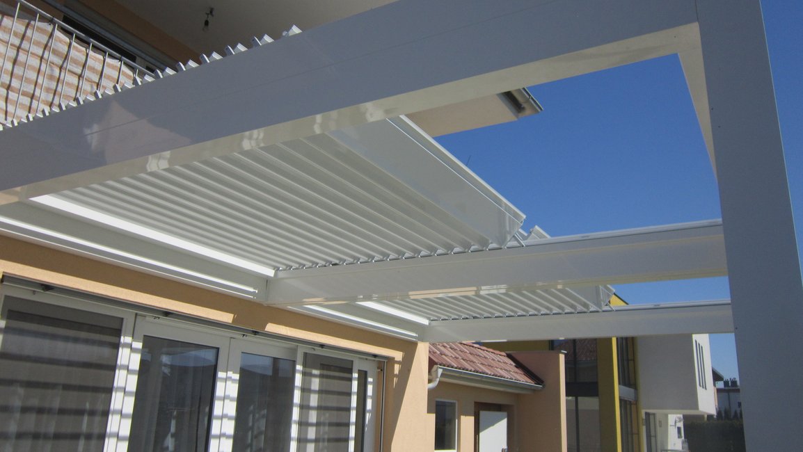Bílá bioklimatická pergola ukotvená k domu, u níž jsou střešní lamely orientovány rovnoběžně s prosklenou fasádou. Na oknech je jasně patrný pruhovaný stín, který tento typ orientace vytváří + alt text: Bioklimatická pergola s lamelami orientovanými rovnoběžně s domem