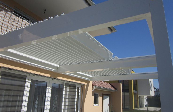 Bílá bioklimatická pergola ukotvená k domu, u níž jsou střešní lamely orientovány rovnoběžně s prosklenou fasádou. Na oknech je jasně patrný pruhovaný stín, který tento typ orientace vytváří + alt text: Bioklimatická pergola s lamelami orientovanými rovnoběžně s domem