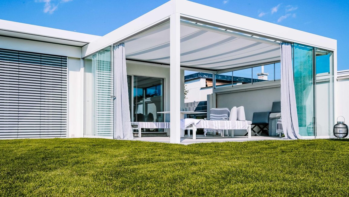 Moderní bílá markýzová pergola s textilní střechou a bočním posuvným zasklením.