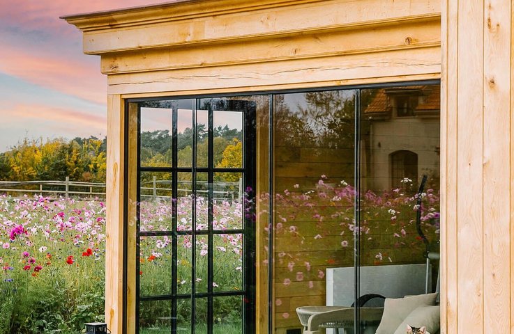 Designový zahradní pavilon z masivního dřeva je uzavřený bezrámovým posuvným zasklením s jemným industriálním dělením skel. Vzniká tak útulný obytný prostor s pohodlným sezením, který zůstává v úzkém kontaktu s okolní květinovou loukou a západem slunce.