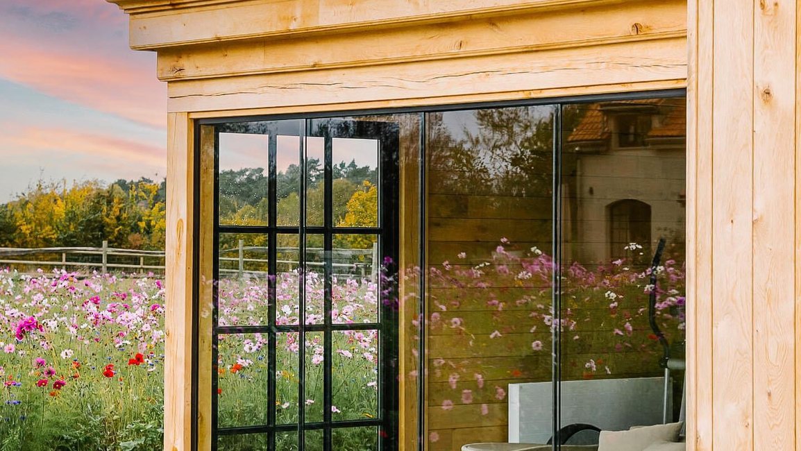 Designový zahradní pavilon z masivního dřeva je uzavřený bezrámovým posuvným zasklením s jemným industriálním dělením skel. Vzniká tak útulný obytný prostor s pohodlným sezením, který zůstává v úzkém kontaktu s okolní květinovou loukou a západem slunce.