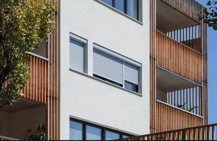 Moderní bytový dům s předokenními roletami v bílé barvě; elegantní kombinace s dřevěným lamelovým obkladem lodžií pro účinné stínění a soukromí.