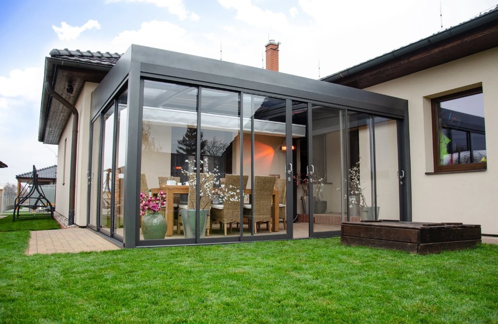 Moderní hliníková pergola u rodinného domu se zasklením po celém obvodu slouží jako komfortní venkovní jídelna. Uvnitř je velký stůl s pohodlnými židlemi, dekorace v květináčích a sálavé topení, takže si majitelé mohou užívat stolování na terase od jara až do podzimu.