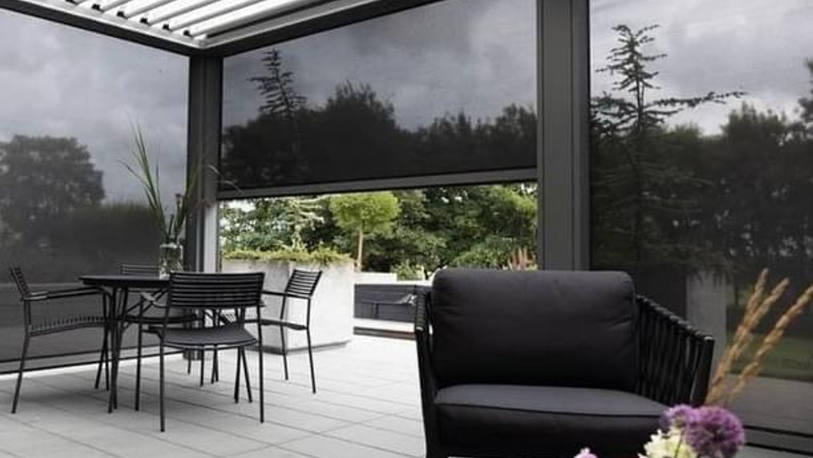 Bioklimatická pergola s hliníkovými lamelami a screenovými roletami, pod nimiž je pohodlné zahradní posezení chráněné před větrem i nízkým sluncem.