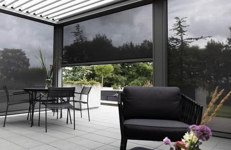 Bioklimatická pergola s hliníkovými lamelami a screenovými roletami, pod nimiž je pohodlné zahradní posezení chráněné před větrem i nízkým sluncem.