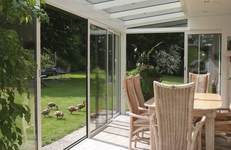 Jídelní kout v zimní zahradě je chráněný rámovým posuvným zasklením a prosklenou střechou. Velké skleněné plochy zajišťují dostatek denního světla a umožňují pohodlný vstup přímo na trávník, takže se interiér přirozeně propojuje se zahradou.