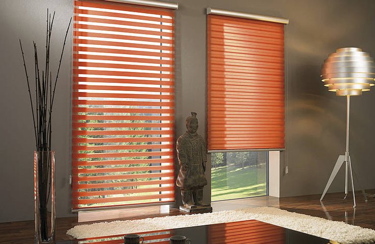 Rolety den a noc v oranžové barvě v moderním obývacím pokoji – jedno okno v režimu průhledných pruhů, druhé v plném zatemnění.