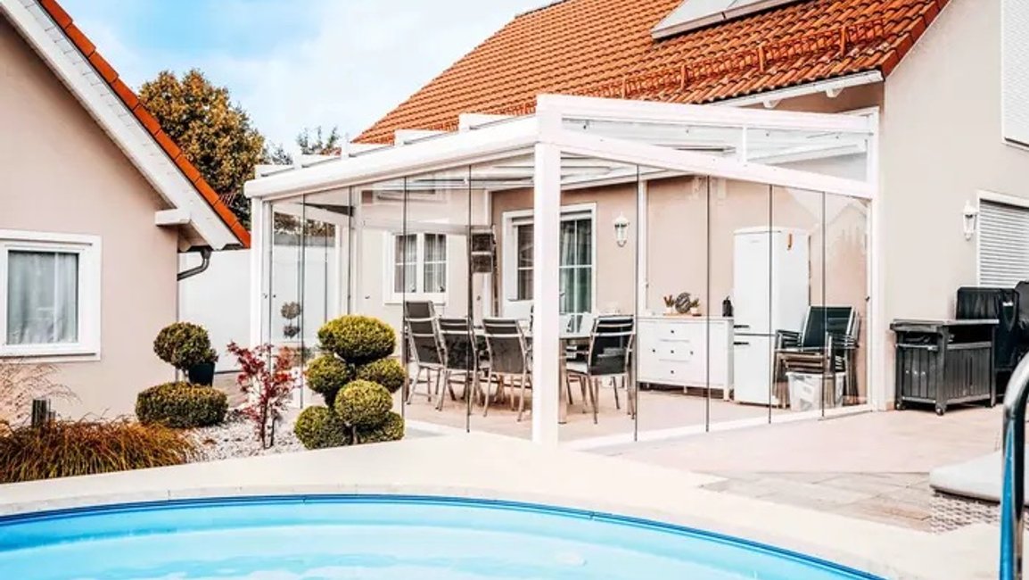 Moderní bílá pergola s bezrámovým zasklením rozšiřuje terasu rodinného domu o chráněné místo pro stolování a odpočinek. Díky proskleným stěnám zůstává výhled na zahradu i bazén otevřený a terasa je využitelná od jara do podzimu.